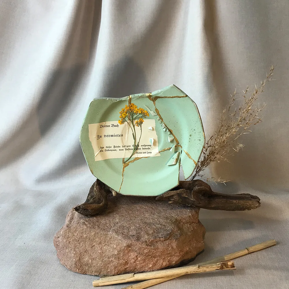Aufrecht stehender, salbeigrüner Kintsugi-Teller auf Treibholz und Sandstein, mit einer Kette aus winzigen Scherben entlang der rechten Goldnaht und einer Buchseite mit der Aufschrift ‚Drittes Buch / Zu vermieten / Romeo und Julia‘.