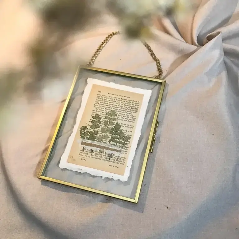 Schräge Draufsicht auf einen rechteckigen Glas-Bilderrahmen mit Goldkante und goldener Gliederkette, darin eine historische Buchseite mit einer grünen Pflanzenillustration, flach arrangiert auf sanft gefaltetem, beigem Leinenstoff mit weicher Vordergrund-Unschärfe von Trockenblumen