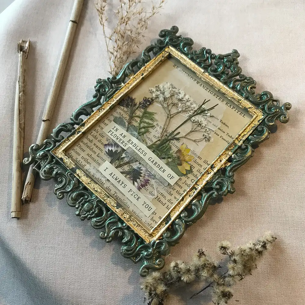 Schräge Draufsicht auf ein gerahmtes Kintsugi- und Wabi-Sabi-Kunstwerk in einem verzierten Barockrahmen in Antik-Grün und Gold, das echte gepresste Wiesenblumen auf einer alten Buchseite in glänzendem Epoxy-Resin konserviert zeigt, platziert auf beigem Stoff neben getrockneten Gräsern.