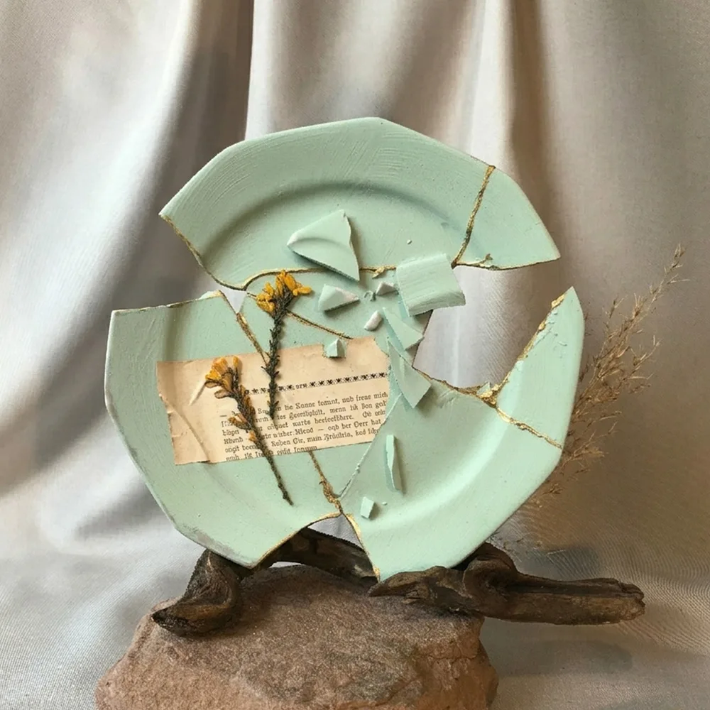 Aufrecht stehender, salbeigrüner Kintsugi-Teller mit sichtbaren Goldnähten, getragen von einem dunklen Treibholz-Ast auf einem rauen Naturstein-Sockel vor beigem Stoff, dekoriert mit einer Buchseite mit dem Textfragment ‚die Sonne scheint‘ und gelben Trockenblumen.