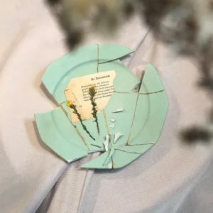 Draufsicht auf einen fragmentierten mintgrünen Kintsugi-Teller mit goldenen Risslinien, kleinen Keramiksplittern, gelben Trockenblumen und einer alten Buchseite auf beigem Leinen.