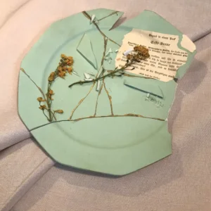 Schrägansicht eines dekonstruierten mintgrünen Kintsugi-Keramiktellers mit plastischen Goldadern, Tonsplittern, feinen grünen Blättern und einer historischen Goethe-Zeittafel auf hellbeigem Stoff