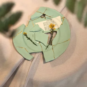 Draufsicht auf einen dekonstruierten mintgrünen Kintsugi-Teller mit filigranen Goldadern, einer historischen Buchseite mit Gedichttext und zarten weißen Blüten auf beigem Leinenstoff.