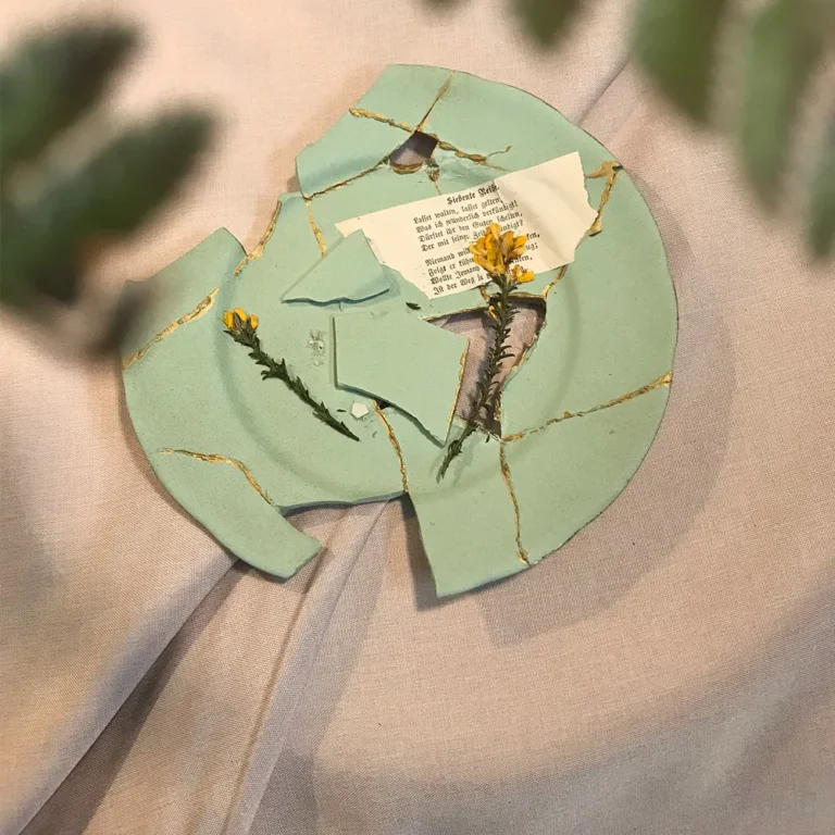 Draufsicht auf einen dekonstruierten mintgrünen Kintsugi-Teller mit filigranen Goldadern, einer historischen Buchseite mit Gedichttext und zarten weißen Blüten auf beigem Leinenstoff.