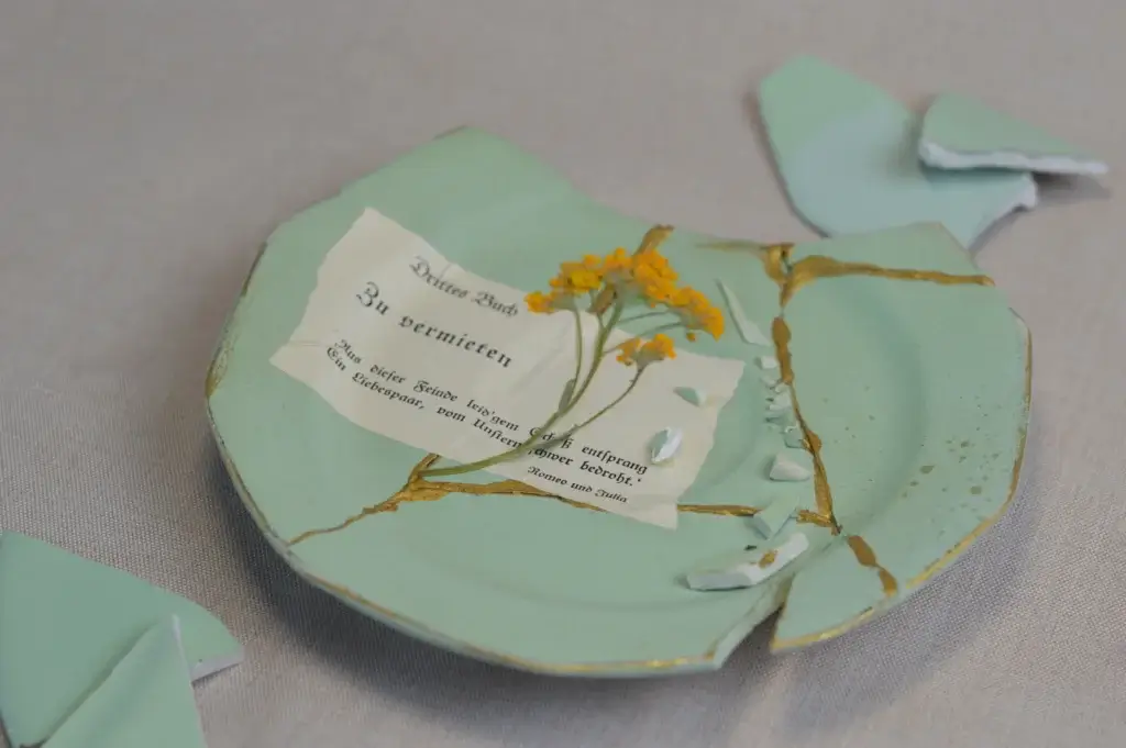 Ein mit goldenem Kintsugi reparierter, hellgrüner Keramikteller auf beigem Leinen, auf dem ein Stück vergilbtes Buchpapier mit altdeutscher Schrift aus Romeo und Julia sowie eine getrocknete gelbe Blüte liegen, umgeben von losen Tonscherben