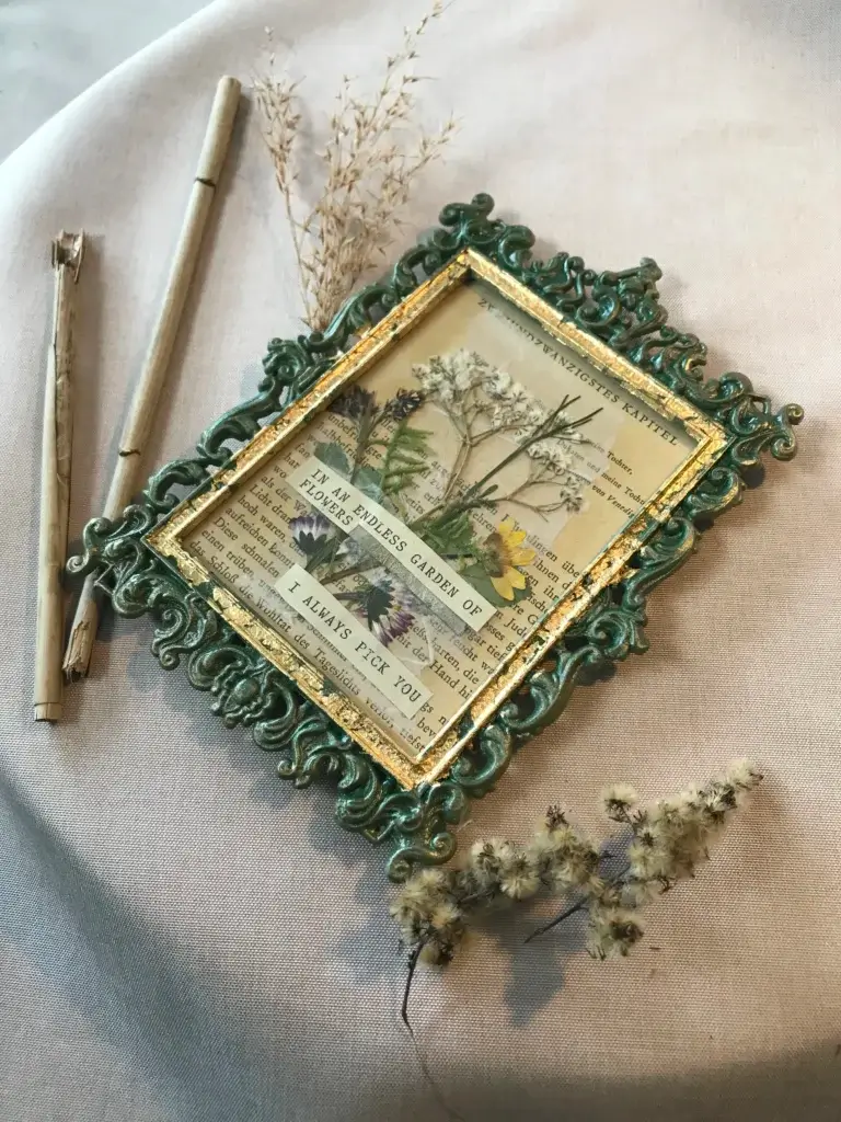 Prunkvoller Barockrahmen in antikem Grün und Gold mit echtem Glas, darin eine historische Buchseite mit der Überschrift ‚ZWEIUNDZWANZIGSTES KAPITEL‘, gepressten Wiesenblumen und der englischen Aufschrift ‚IN AN ENDLESS GARDEN OF FLOWERS I ALWAYS PICK YOU‘, arrangiert auf beigem Leinen