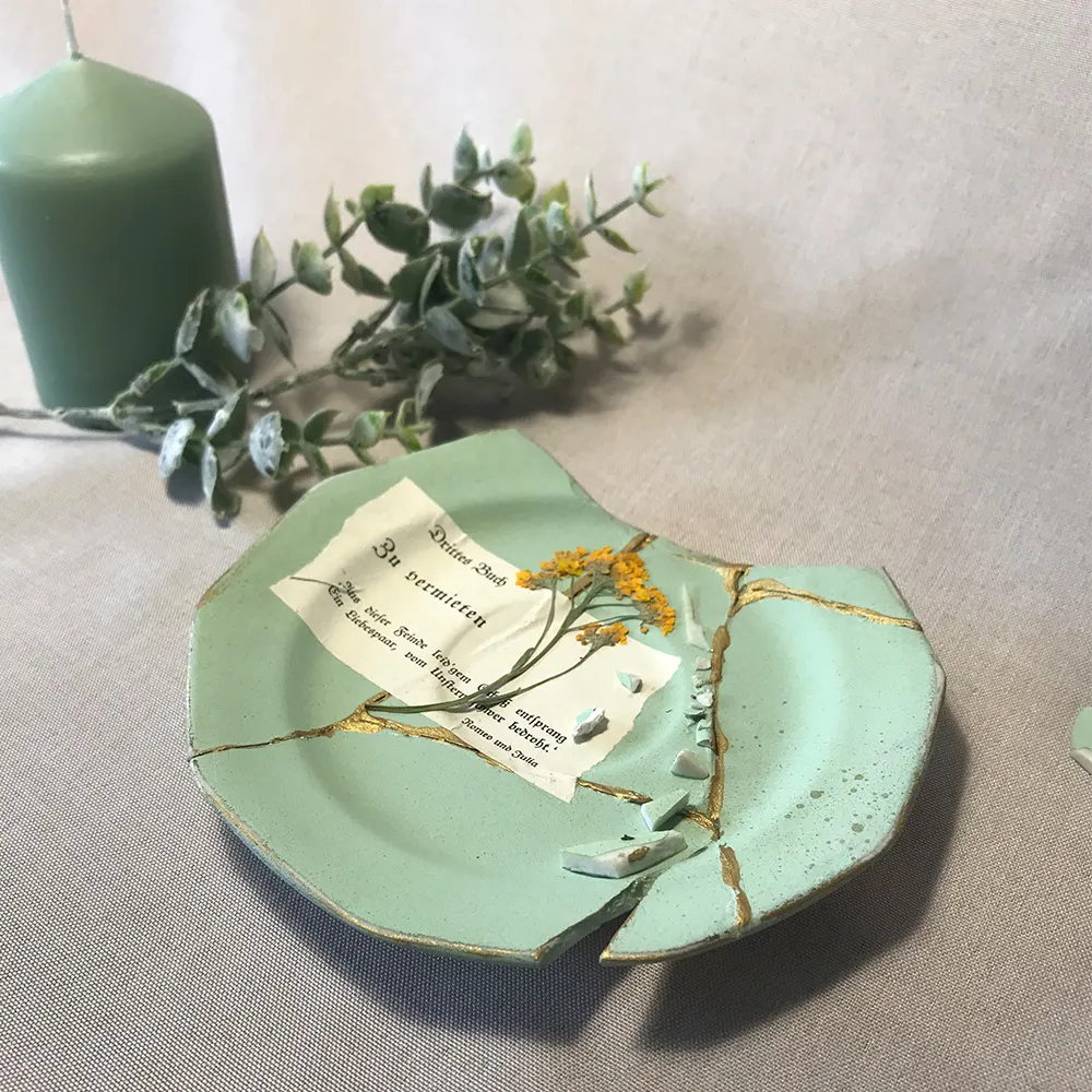 Schräge Draufsicht auf einen hellgrünen Kintsugi-Teller mit goldenen Linien auf grauem Leinenstoff, im Hintergrund eine salbeigrüne Stumpenkerze und ein Eukalyptuszweig, dekoriert mit einer Buchseite mit der Aufschrift ‚Zu vermieten