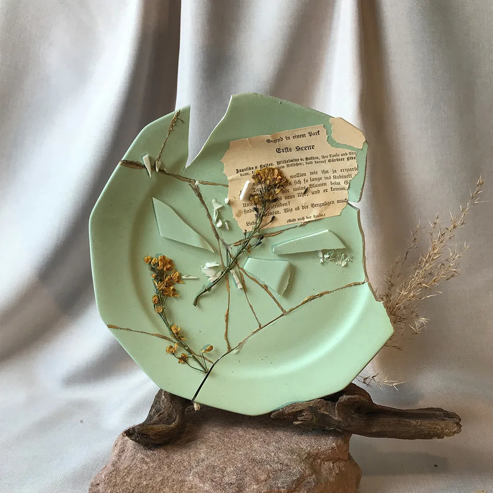 Kintsugi-Skulptur von Lineativ Studio: Ein mintgrüner, fragmentierter Keramikteller mit Goldnähten, antikem Buchpapier und einer gelben Blüte, arrangiert auf einem Sockel aus Naturstein und Treibholz im Wabi-Sabi-Stil als hochwertiges Geschenk für Kintsugi Liebhaber.
