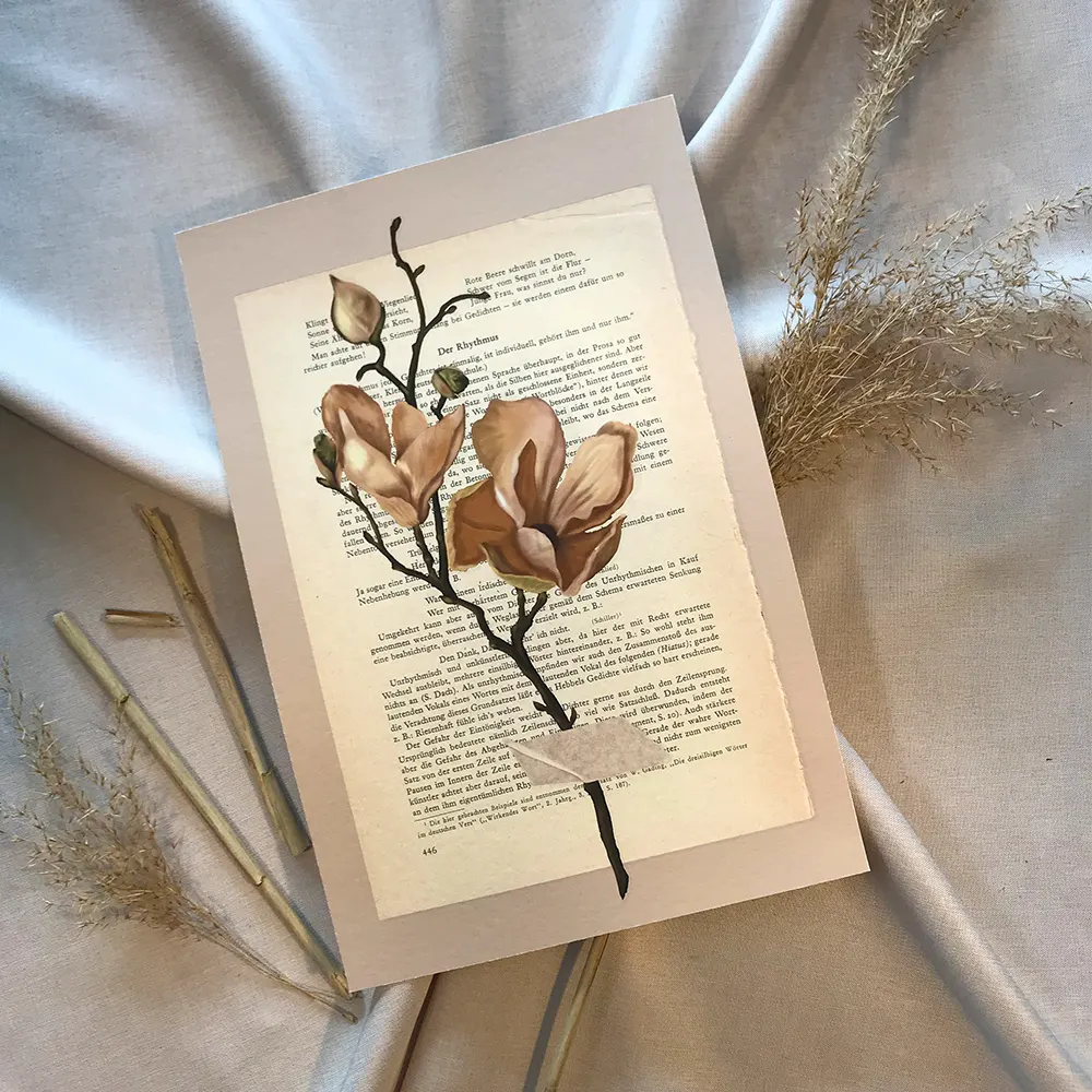 Schräge Vogelperspektive auf einen Magnolien-Kunstdruck auf vergilbter Buchseite, flach arrangiert auf sanft gefaltetem, grau-beigen Leinenstoff, umgeben von echten, filigranen Trockengräsern mit ausgeprägtem Schattenwurf.