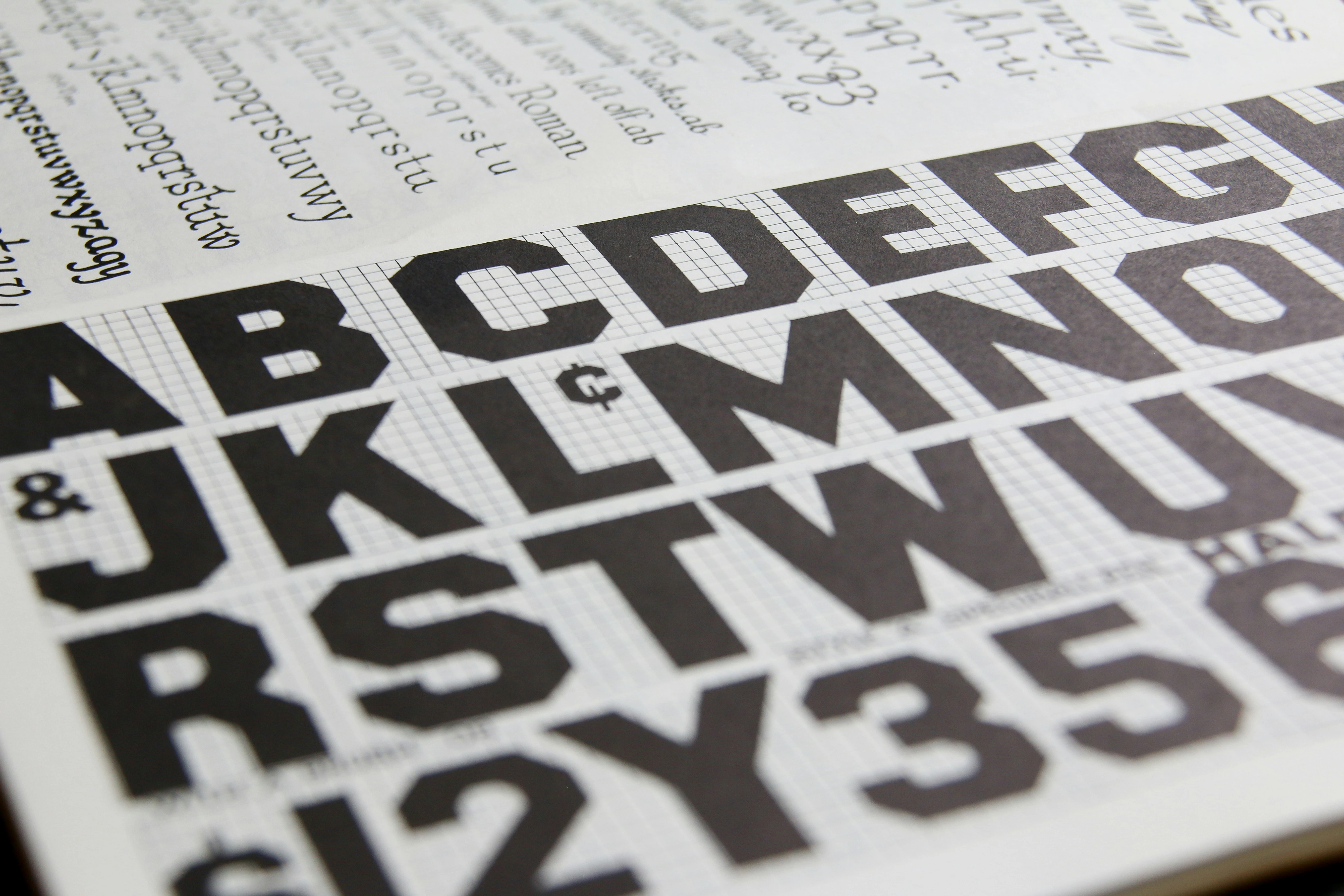 Nahaufnahme eines Typografie-Musterbuchs mit serifenlosen, fetten Großbuchstaben des Alphabets im Vordergrund und feiner Kalligrafie im Hintergrund.
