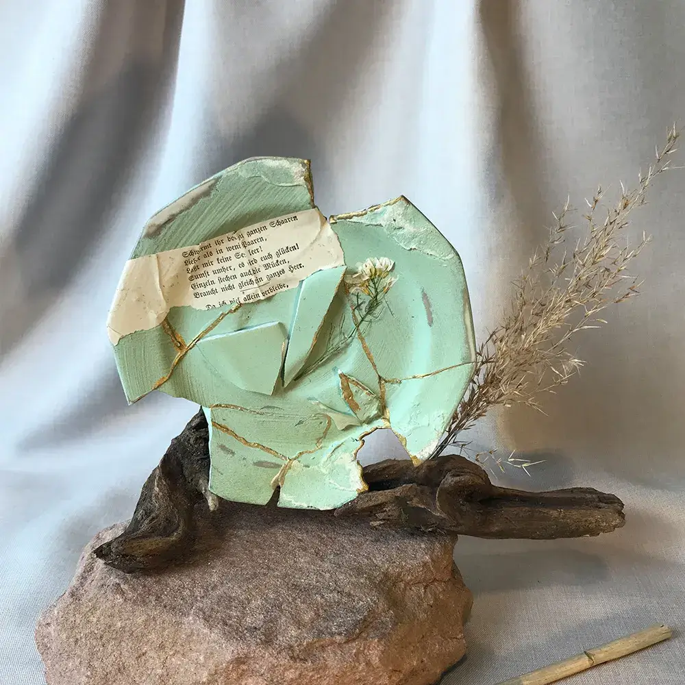 Aufrecht stehender, zerbrochener Kintsugi-Teller in Salbeigrün mit goldenen Rissen, montiert auf einem verwitterten Treibholz-Ast auf einem rauen Naturstein, dekoriert mit einer alten Buchseite in Frakturschrift und kleinen gelben Trockenblumen.
