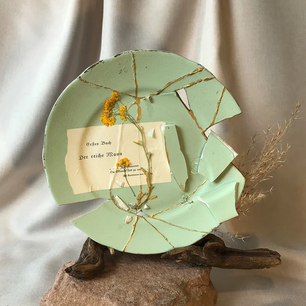 Aufrecht stehender, salbeigrüner Kintsugi-Teller auf Treibholz und Sandstein, mit einem großen keilförmigen Ausbruch rechts, einem vertikalen Scherbenfragment im Zentrum und einer Buchseite mit dem Text ‚Erstes Buch / Der reiche Mann‘.