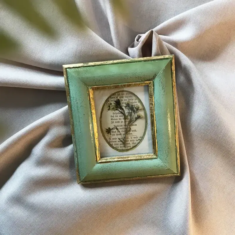 Draufsicht auf ein gerahmtes Wabi-Sabi-Kunstwerk mit getrockneten Blüten auf einer alten Buchseite in einem mintgrünen Vintage-Rahmen mit Goldrand, liegend auf elegant gefaltetem, beigem Stoff.