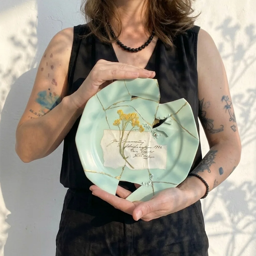 Kintsugi-Teller in Mintgrün mit echtem Gold repariert, verziert mit gepressten gelben Blüten und einer antiken Buchseite, gehalten von einer Künstlerin im Lineativ Studio.