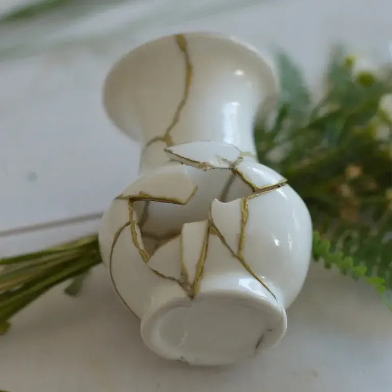 Eine weiße Kintsugi-Keramikvase liegt leicht nach vorne gekippt auf einer hellen Oberfläche, fotografiert aus der Vogelperspektive. Goldene Risse verbinden die Scherben, und im Bauch der Vase klafft ein großes Loch, das den Blick ins leere Innere freigibt. Im Hintergrund liegen unscharf grüne Farnzweige.