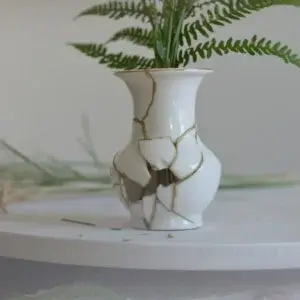 Eine kleine, weiße Keramikvase steht auf einer hellen Oberfläche und hält grüne Farnzweige. Die Vase war zerbrochen und wurde im Stil der japanischen Kintsugi-Kunst mit goldenen Rissen wieder zusammengesetzt, wobei einige Scherben im Bauch der Vase fehlen und ein Loch hinterlassen.