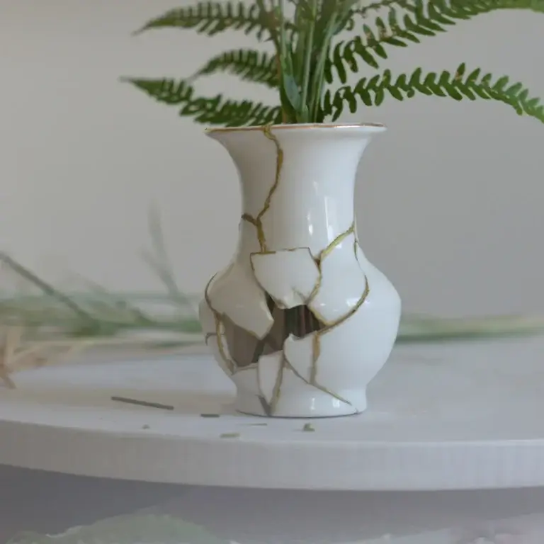 Eine kleine, weiße Keramikvase steht auf einer hellen Oberfläche und hält grüne Farnzweige. Die Vase war zerbrochen und wurde im Stil der japanischen Kintsugi-Kunst mit goldenen Rissen wieder zusammengesetzt, wobei einige Scherben im Bauch der Vase fehlen und ein Loch hinterlassen.