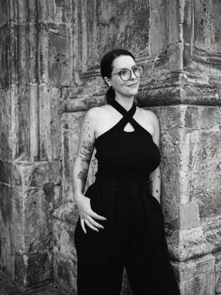 Schwarz-Weiß-Portrait einer lächelnden Frau mit Brille, Tätowierungen am Arm und hochgestecktem Haar. Sie trägt ein schwarzes Neckholder-Top mit gekreuzten Trägern und eine dunkle Hose, während sie sich an eine historische, strukturierte Steinwand lehnt.