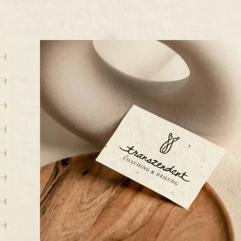Eine strukturierte Visitenkarte aus handgeschöpftem Papier mit Einschlüssen liegt auf einem runden Holzteller. Auf der Karte ist das geschwungene Logo „transzendent – COACHING & HEILUNG“ des Lineativ Studios aus Hauenstein zu sehen. Im Hintergrund befindet sich eine minimalistische, beige Keramikskulptur.