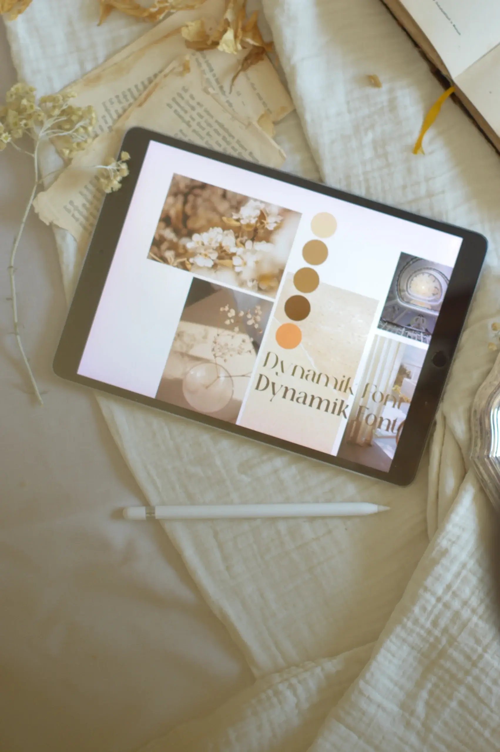 Ein Tablet zeigt ein ästhetisches Moodboard mit hellen Blütenfotos, einer Farbpalette in Beige-, Braun- und Orangetönen sowie dem Schriftzug „Dynamik Font“. Das Tablet liegt schräg auf einer hellen Decke, umgeben von einem Eingabestift, vergilbten Buchseiten und getrockneten Blumen.