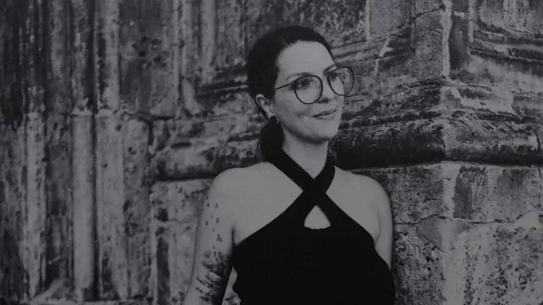 Schwarz-Weiß-Portrait einer lächelnden Frau mit Brille, Tätowierungen am Arm und hochgestecktem Haar. Sie trägt ein schwarzes Neckholder-Top mit gekreuzten Trägern und eine dunkle Hose, während sie sich an eine historische, strukturierte Steinwand lehnt.