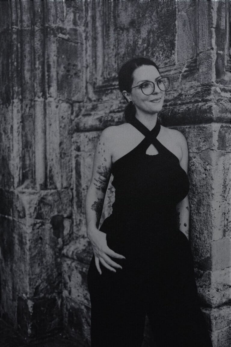 Schwarz-Weiß-Portrait einer lächelnden Frau mit Brille, Tätowierungen am Arm und hochgestecktem Haar. Sie trägt ein schwarzes Neckholder-Top mit gekreuzten Trägern und eine dunkle Hose, während sie sich an eine historische, strukturierte Steinwand lehnt.