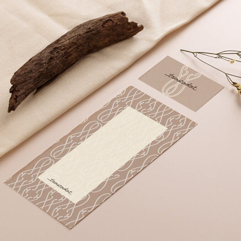 Ein Marken-Mockup von Lineativ Studio für die Marke „transzendent“. Auf einem beigen Untergrund liegen diagonal angeordnet eine Visitenkarte und ein länglicher Papierbogen im erdigen Farbton Mocha Mouse. Beide zeigen ein elegantes, weißes Linienmuster aus geschwungenen Ornamenten und den Markennamen in feiner Schreibschrift. Links oben liegt ein verwittertes Stück Holz, rechts ragen zarte Trockenblumen ins Bild.