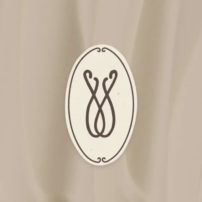Das offizielle Marken-Signet von „transzendent“ präsentiert auf einem elegant fallenden Stoffhintergrund im Farbton Mocha Mouse. Das Logo ist als hochkant ausgerichtetes Oval in Papyrus-Weiß gestaltet, umrahmt von einer feinen dunklen Linie mit kleinen Zierschnörkeln oben und unten. Im Zentrum des Ovals befindet sich das abstrakte, geschwungene Monogramm im tiefen Farbton Plum Nior, das an ineinander verschlungene Linien oder ein florales Element erinnert.