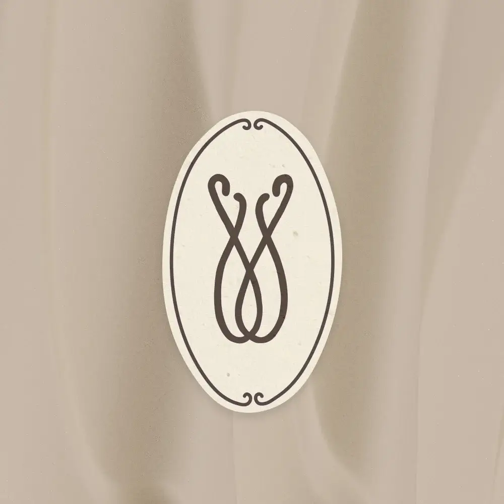 Das offizielle Marken-Signet von „transzendent“ präsentiert auf einem elegant fallenden Stoffhintergrund im Farbton Mocha Mouse. Das Logo ist als hochkant ausgerichtetes Oval in Papyrus-Weiß gestaltet, umrahmt von einer feinen dunklen Linie mit kleinen Zierschnörkeln oben und unten. Im Zentrum des Ovals befindet sich das abstrakte, geschwungene Monogramm im tiefen Farbton Plum Nior, das an ineinander verschlungene Linien oder ein florales Element erinnert.