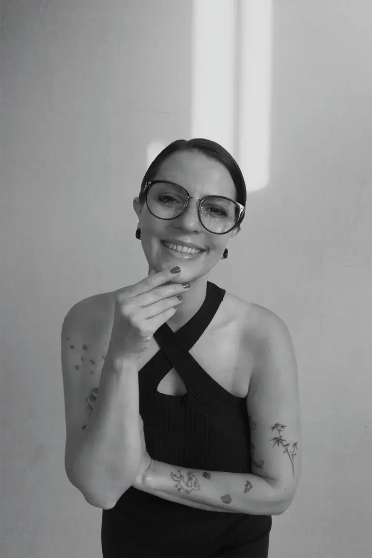 Ein ästhetisches Schwarz-Weiß-Portrait von Jennifer Maier, Brand Designerin für holistische Markenführung bei Lineativ Studio. Sie blickt lächelnd in die Kamera, trägt eine markante, runde Brille, Ohrringe und ein dunkles Neckholder-Top mit gekreuzten Trägern. Ihre Hand berührt sanft ihr Kinn, und auf ihren Armen sind filigrane Tattoos zu sehen. Der Hintergrund ist eine minimalistische, helle Wand mit einem sanften Lichtstrahl.